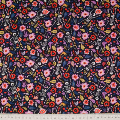 Pink flowers and happy, dancing skeletons are printed on an indigo 100% cotton, halloween fabric with a cm ruler at the bottom