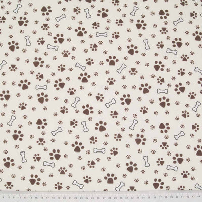Small muddy brown dog paw prints and white bones are printed on a cream polycotton fabric with a cm ruler at the bottom