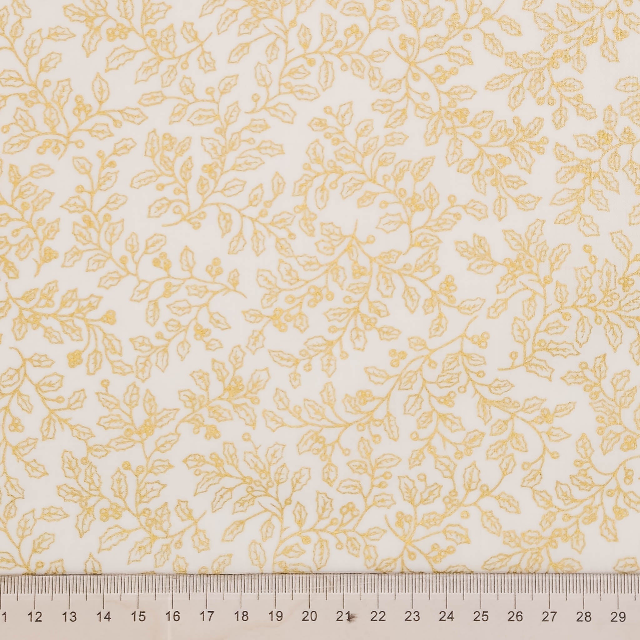A close-up of a fabric or wallpaper pattern featuring a delicate yellow-gold leafy vine design on a warm off-white background, with a ruler at the bottom showing measurement scale.