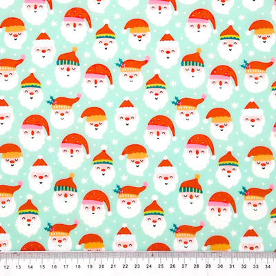 Pattern of cartoon Santa Claus faces with colorful hats on a light blue polycotton fabric, with a ruler at the bottom.