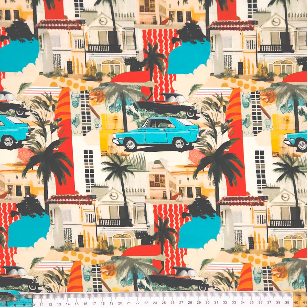A cool graphic design featuring a vintage blue car traveling amongst palm trees, holiday houses and bold areas of stripy red and blue patchwork, digitally printed on a cotton jersey fabric with a cm ruler