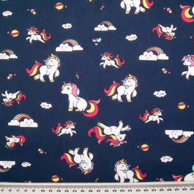 Cute white unicorns, colourful rainbows and stars are printed on a navy polycotton fabric with a ruler at bottom for size perspective