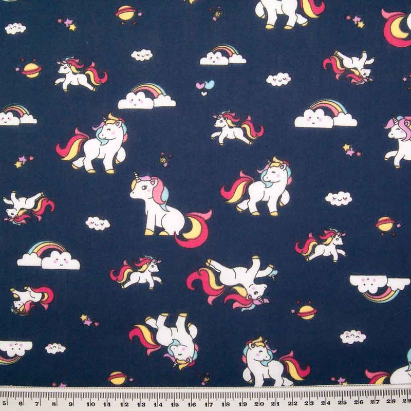 Cute white unicorns, colourful rainbows and stars are printed on a navy polycotton fabric with a ruler at bottom for size perspective