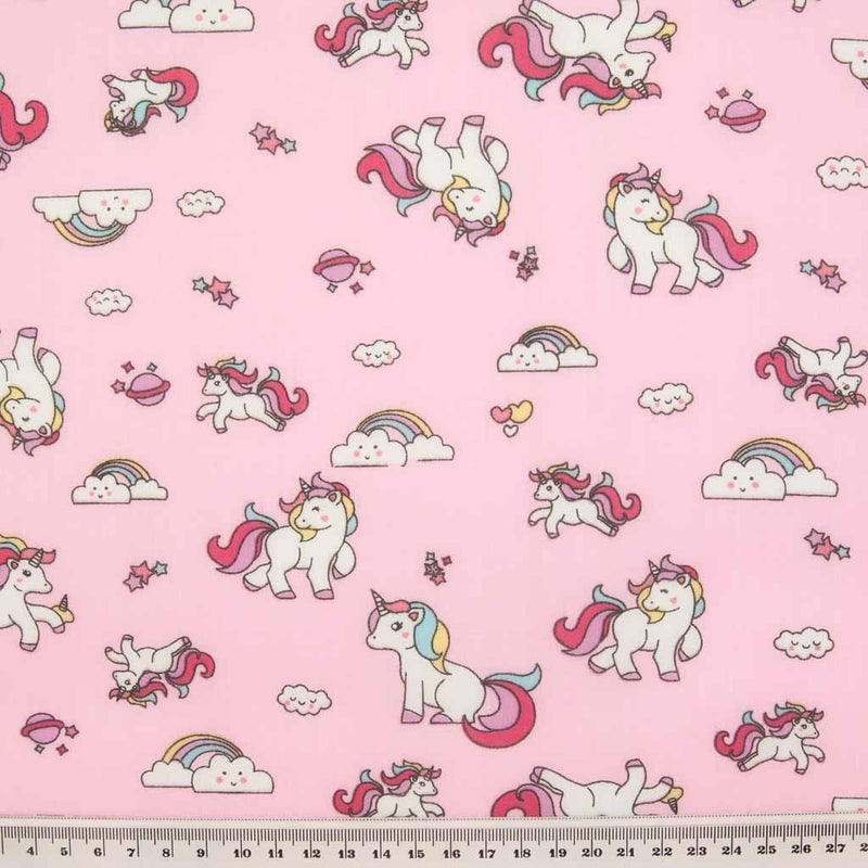 Cute white unicorns, colourful rainbows and stars are printed on a pink polycotton fabric with a ruler at the bottom for size perspective