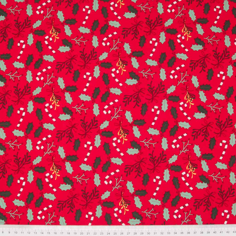 Festive green holly sprigs with candy canes are printed on a red christmas cotton fabric with a cm ruler at the bottom