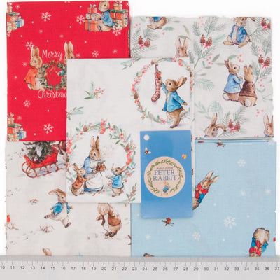 Five cotton fat quarters arranged in a square with a red, a blue and three white backgrounds and festive Peter Rabbit scenes printed on each.. With a cm ruler at the bottom