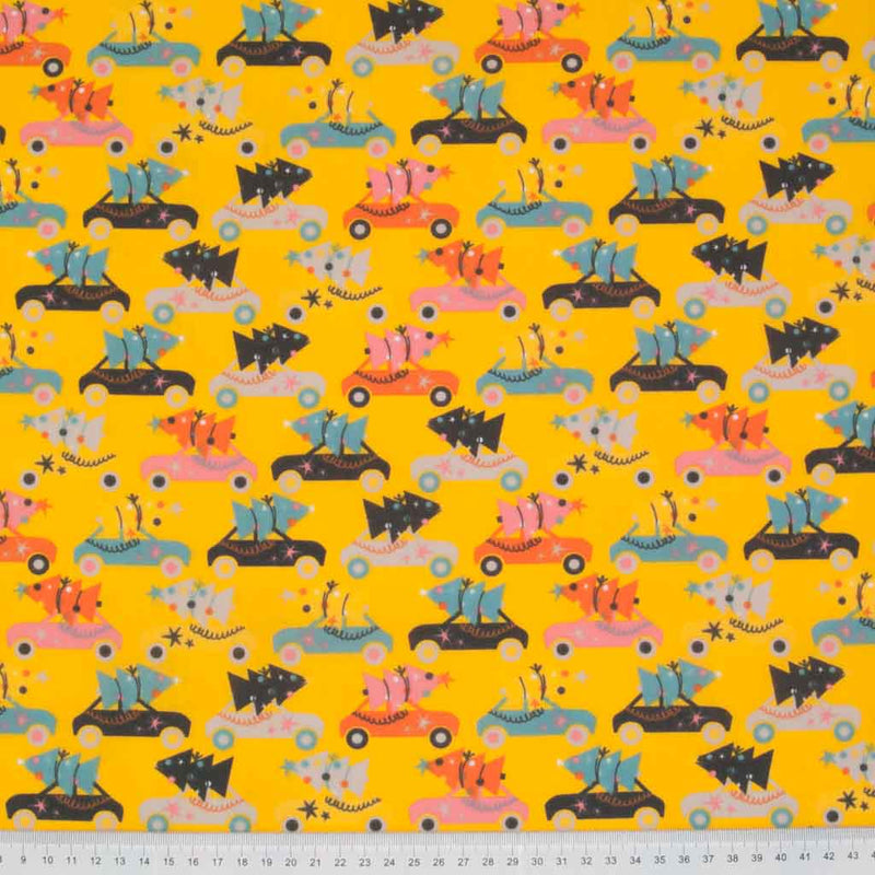 Rows of colourful cars carrying christmas trees are printed on a yellow polycotton fabric with a cm ruler at the bottom