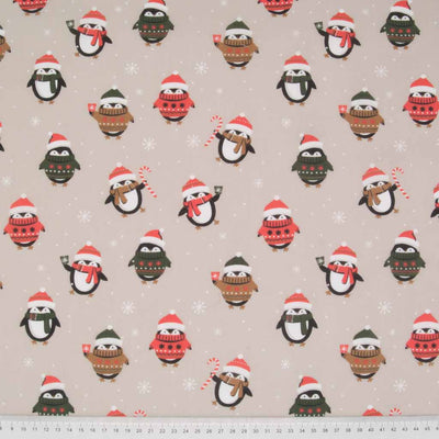 Festive penguins wearing christmas jumpers and wooly hats are printed on a beige polycotton fabric with a cm ruler at the bottom