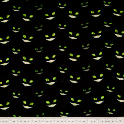 Green grinning cheshire cat faces with glowing eyes are printed on a black, 100% cotton halloween fabric with a cm ruler at the bottom