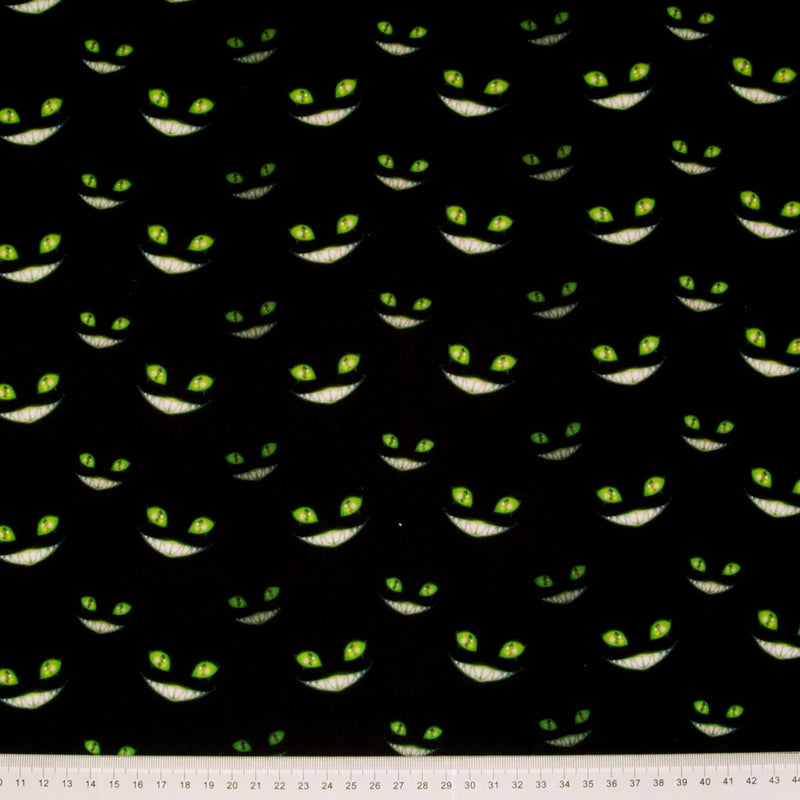 Green grinning cheshire cat faces with glowing eyes are printed on a black, 100% cotton halloween fabric with a cm ruler at the bottom