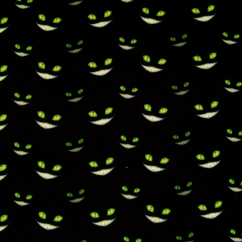 Green grinning cheshire cat faces with glowing eyes are printed on a black, 100% cotton halloween fabric