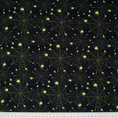Bright neon green spiders on cobwebs are printed on a black, 100% cotton halloween fabric with a cm ruler at the bottom
