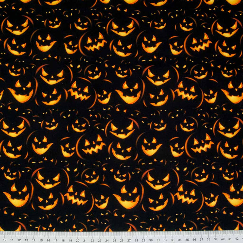 Menacing pumpkin faces in glowing orange are printed on a black 100% cotton halloween fabric with a cm ruler at the bottom