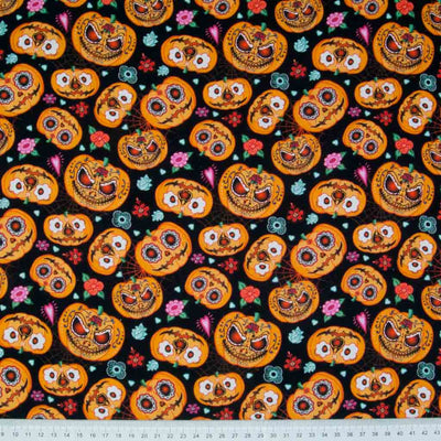 Orange halloween pumpkins with a day of the dead floral design are printed on a black, 100% cotton fabric with a cm ruler at the bottom