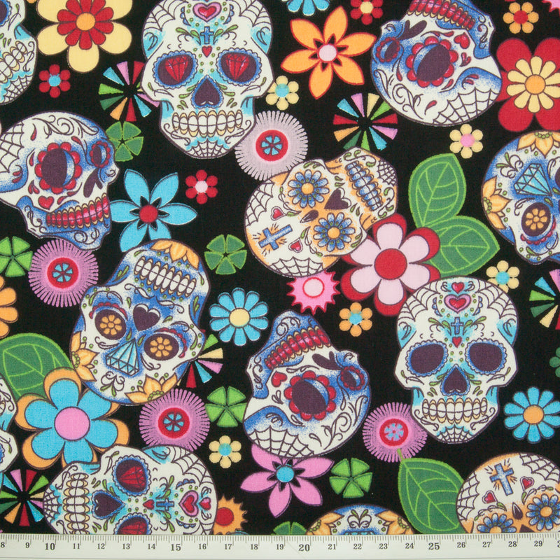 A Rose & Hubble cotton fabric with bright and colourful skulls and flowers printed on a black background with ruler for size perspective