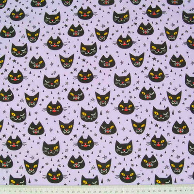 Halloween polycotton fabric printed with winking black cats on a lilac background with a cm ruler at the bottom