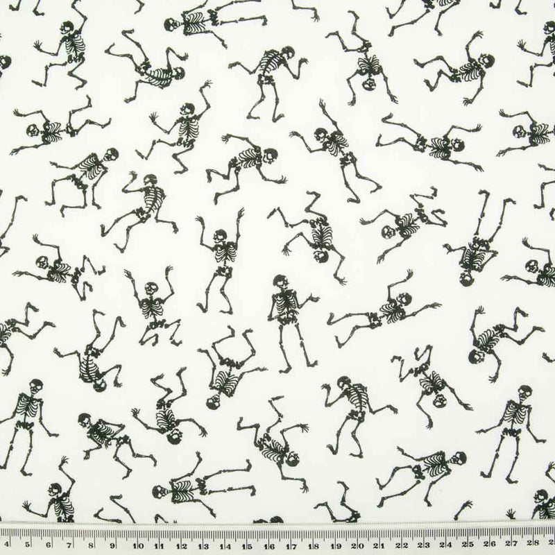 Black mini skeletons in a scattered pattern dancing on white polycotton halloween fabric with a cm ruler at the bottom