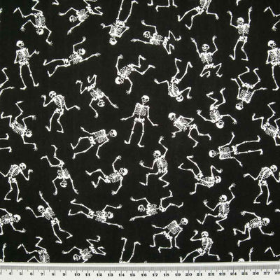 White mini skeletons in a scattered pattern dancing on black polycotton halloween fabric with a cm ruler at the bottom