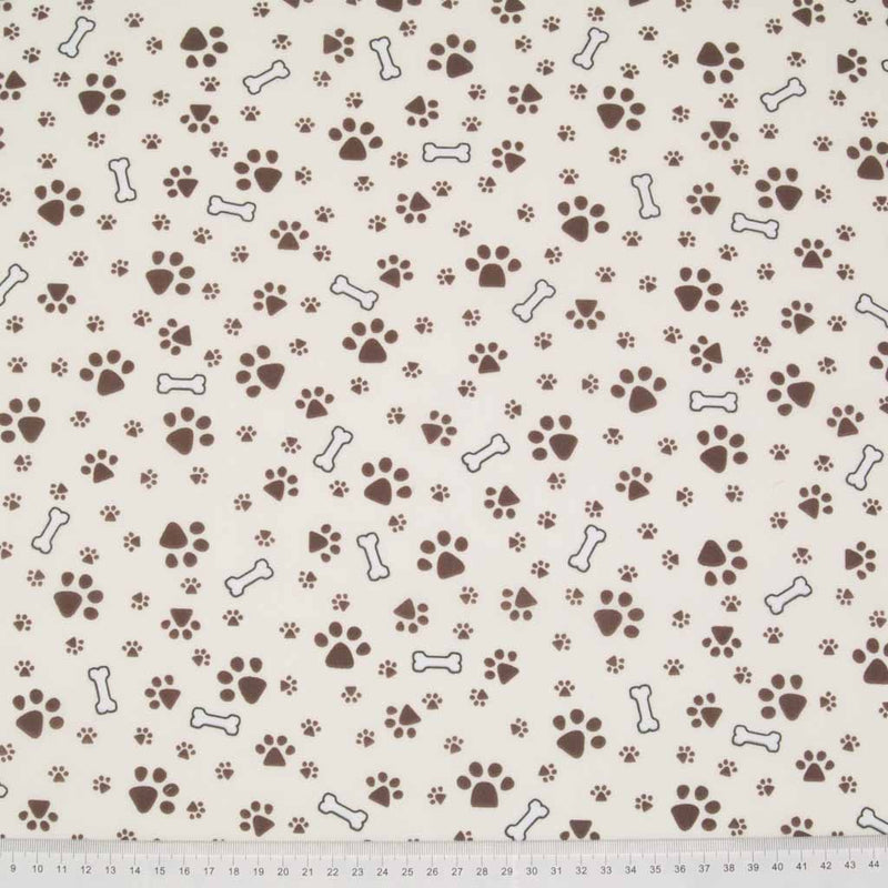 Small muddy brown dog paw prints and white bones are printed on a cream polycotton fabric with a cm ruler at the bottom