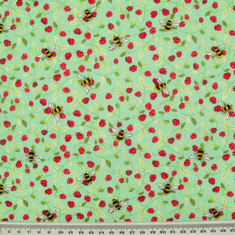 Small red strawberries and yellow bees are printed on a meadow green cotton poplin fabric by Rose & Hubble with a cm ruler at the bottom