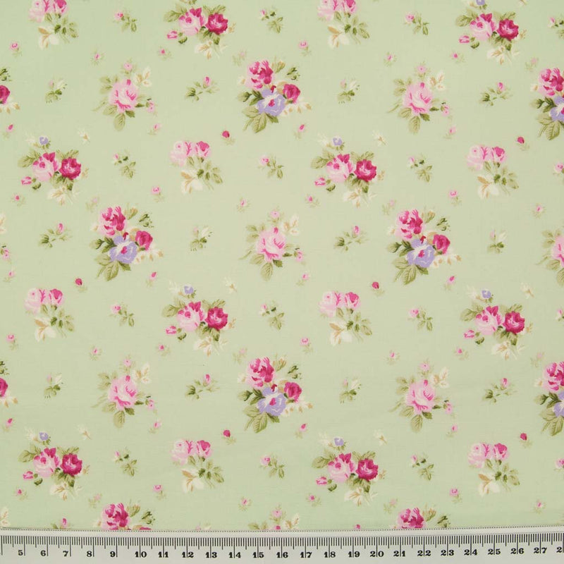 Small bouquets of pink roses are printed on a meadow green cotton poplin fabric with a cm ruler at the bottom