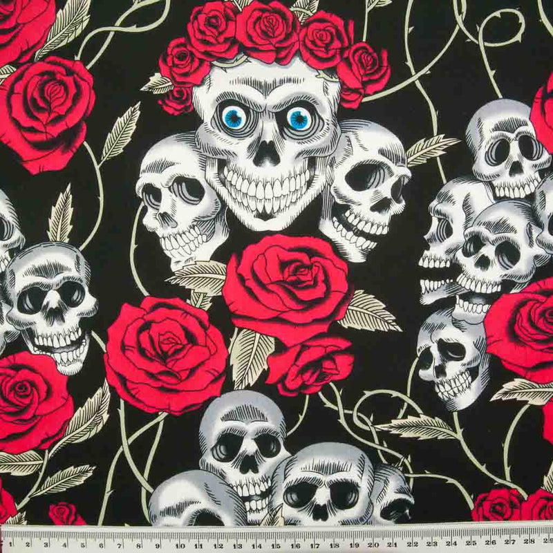Gothic white skulls with blue eyes crowned with bright red roses are printed on a black 100% cotton poplin fabric by Rose and Hubble with a ruler for size perspective