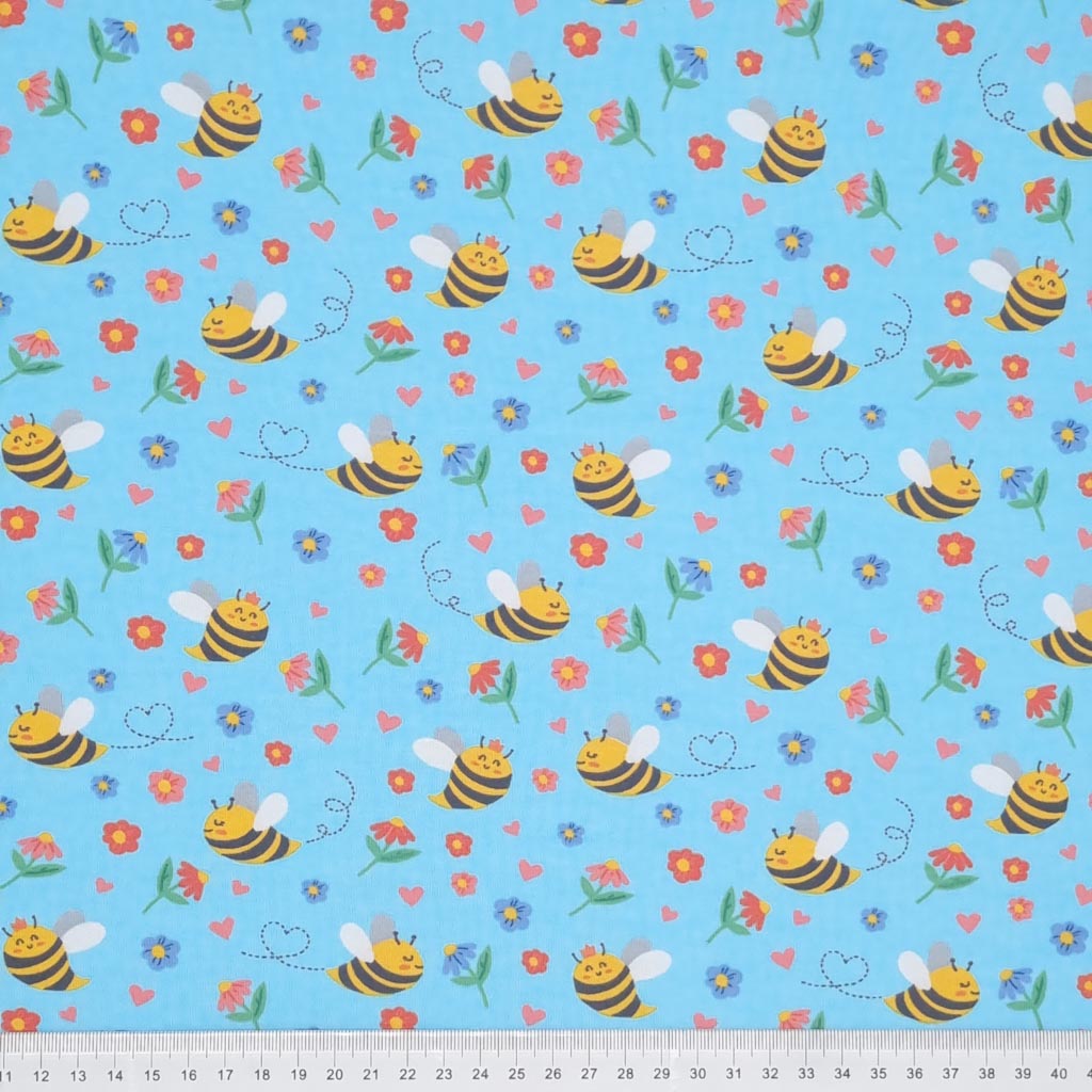 Smiling bees, flowers and hearts printed on a sky blue cotton jersey fabric with a cm ruler at the bottom