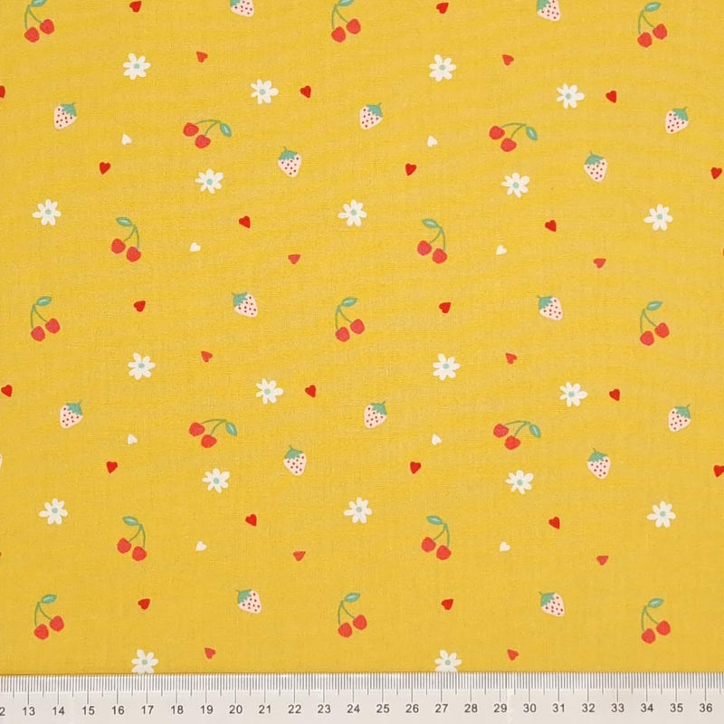 Cherries, strawberries, flowers and hearts are printed on an ochre coloured cotton poplin with a cm ruler at the bottom