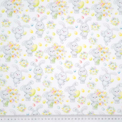 Cute easter bunnies holding spring flowers are printed on a white cotton fabric with a cm ruler at the bottom