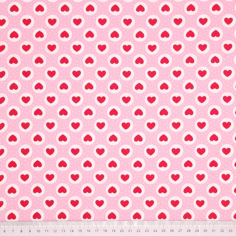 Red hearts and small white spots are printed on a pink cotton poplin fabric by Rose & Hubble with a cm ruler at the bottom