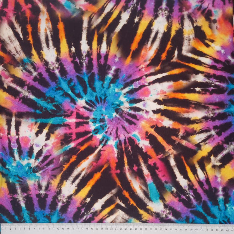 A brightly coloured tie dye viscose fabric printed with a spiralling catherine wheel design with a cm ruler at the bottom